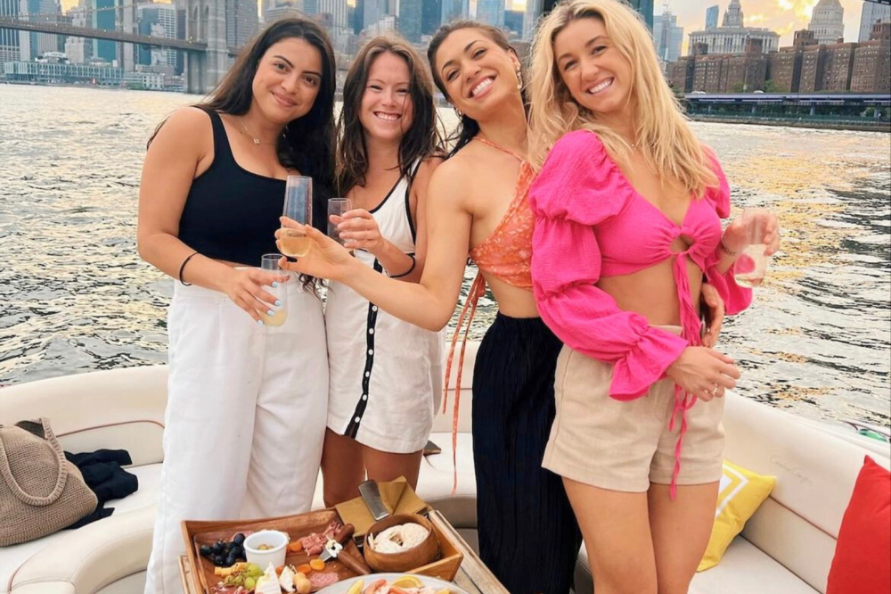 Four women on a boat birthday party with drinks, NYC in the background, charcuterie board on table.