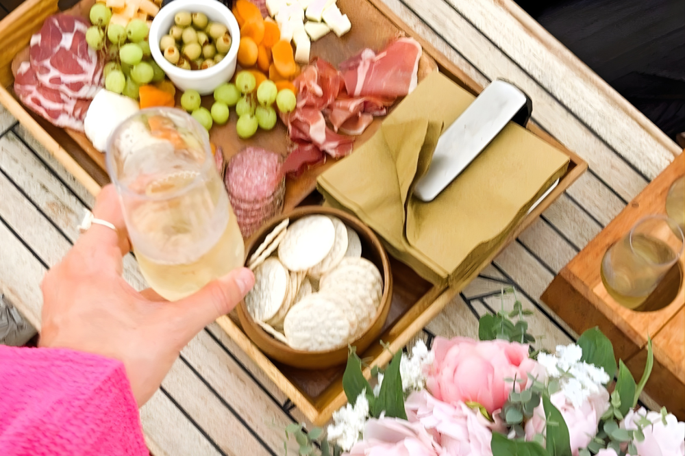 Hand holding drink over charcuterie board and boat catering with flowers on a wooden table in New York.