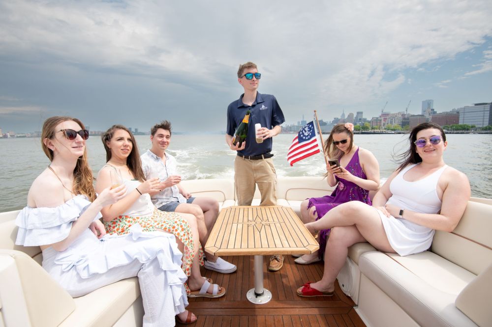 Six people on a boat charter, one holding a champagne bottle, others chatting and drinking, NYC skyline in the background.
