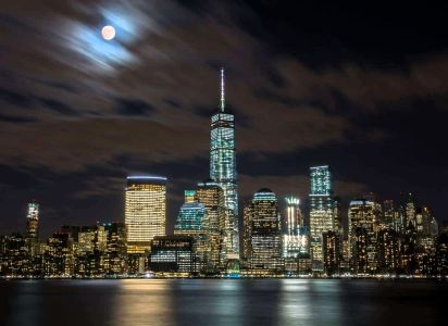 Night view of NYC skyline with illuminated skyscrapers and a full moon during a night boat tour.