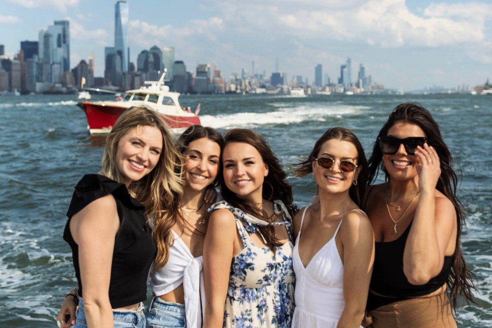 Five women smiling on a private boat tour of New York with the city skyline and a red boat in the background.