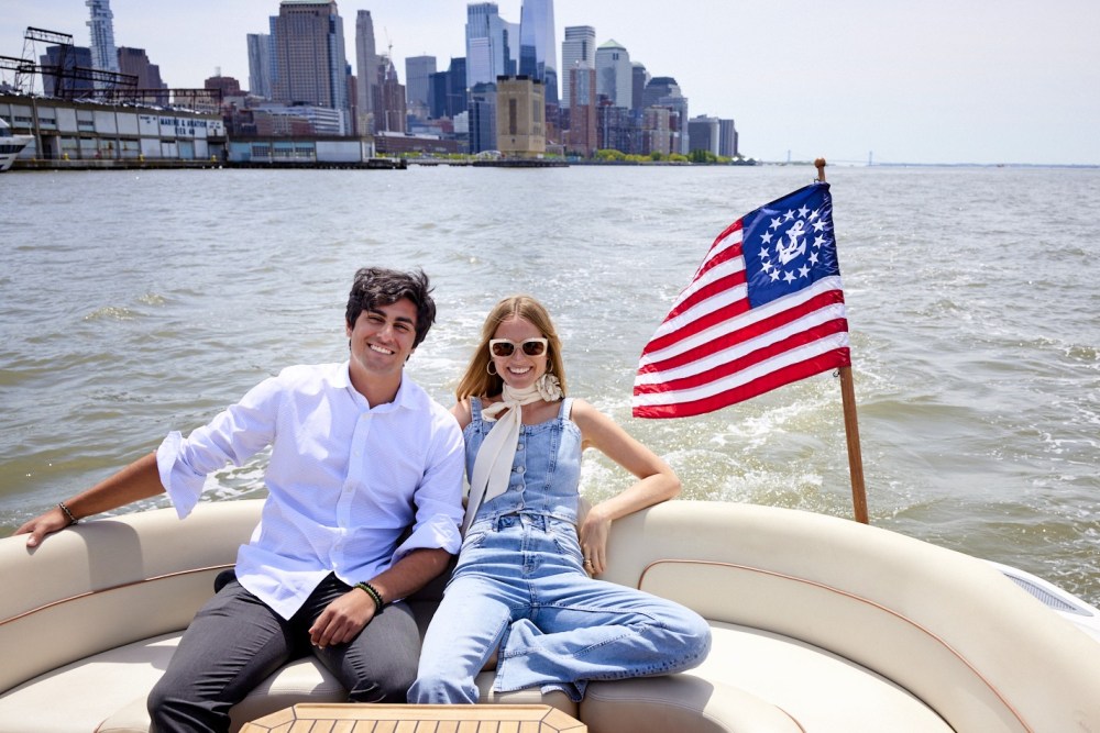 Two people smiling on a private yacht charter with a historical American flag, NYC skyline in the background.