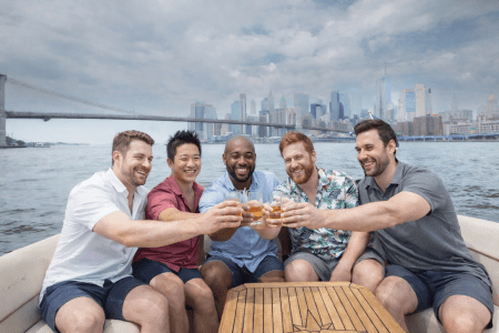 Five men toasting with drinks on a boat with a city skyline in the background.