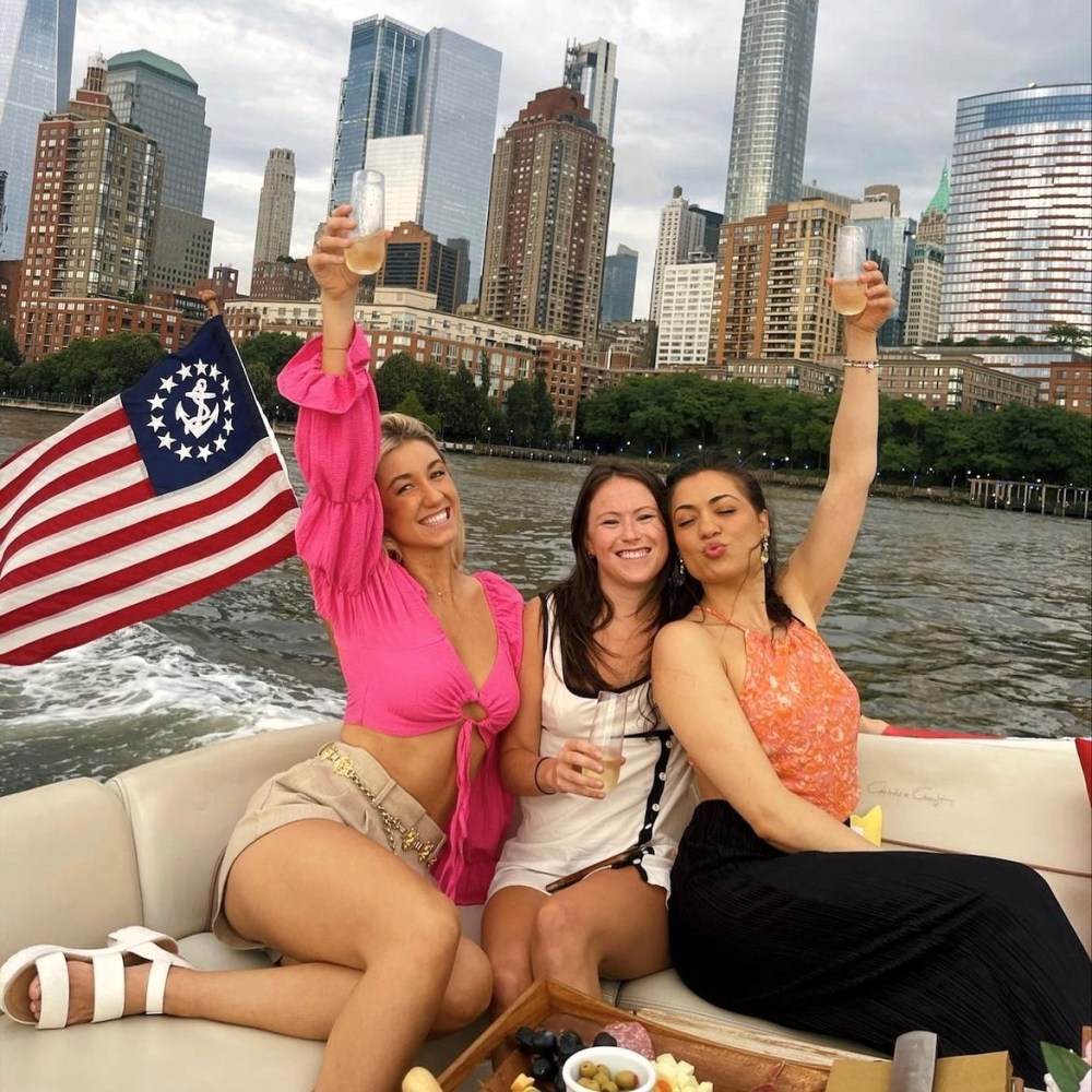 Three women on a yacht rental with drinks, NYC skyline and flag in background.