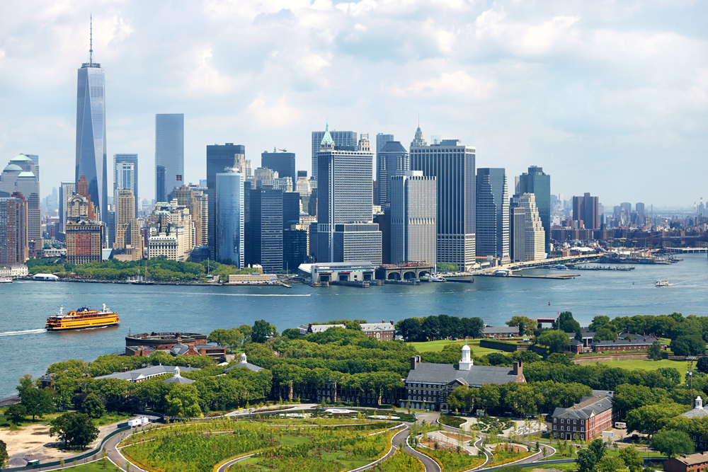 Skyline of Manhattan with skyscrapers, river, ferry, and park in foreground on a sunny day.