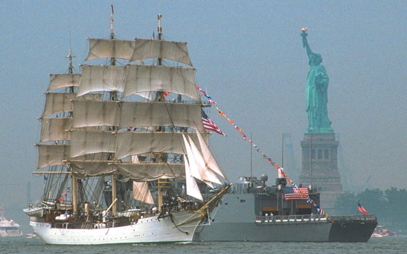 Tall ship and naval vessel near Statue of Liberty with flags in the NY Harbor.